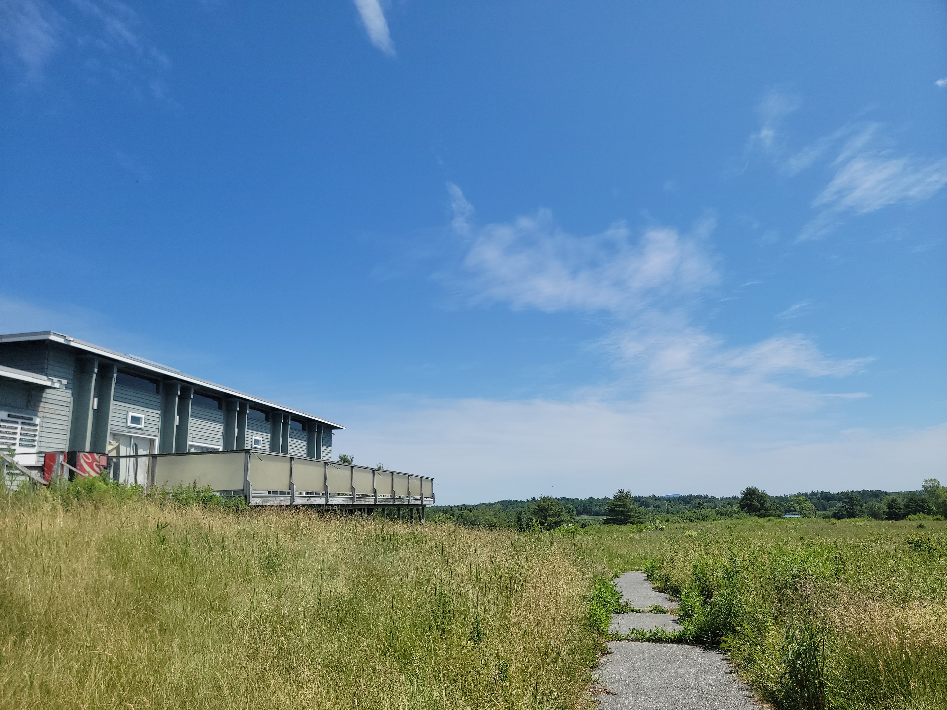 a path leads through tall grass toward blue sky and an old wooden building with windows overlooking the bay